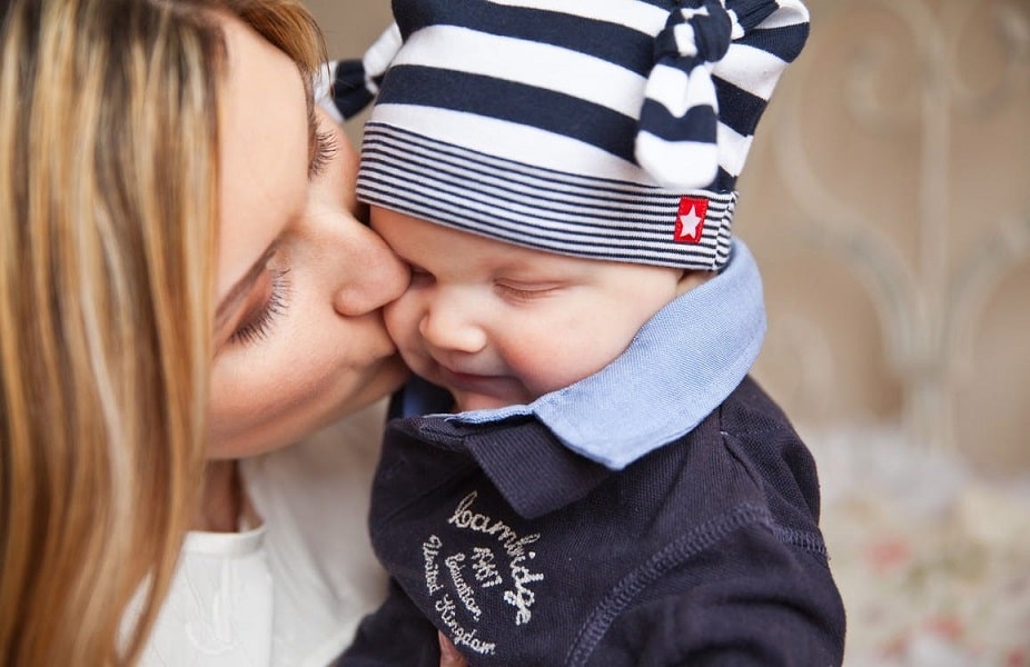 A new mother fondly kissing her baby boy dressed in a blue shirt and cap.